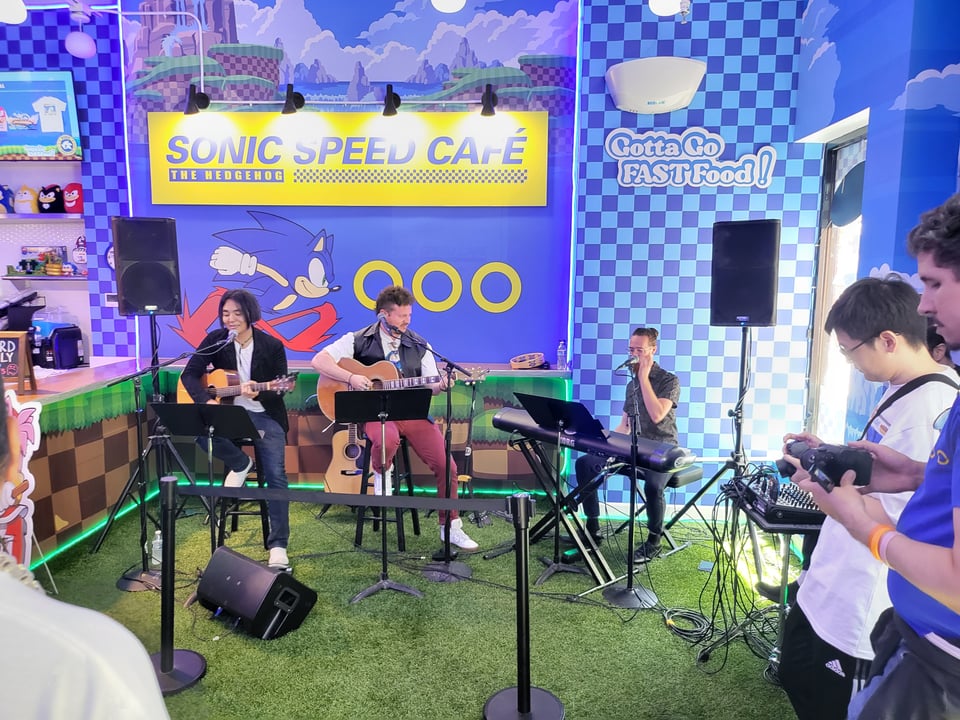 Three musicians--two on guitar and one on keyboard--play music from Sonic the Hedgehog in the Sonic Speed Cafe restaurant. They're set up in one corner under a picture of Sonic running for rings, a blue checkerboard pattern, and the slogan "Gotta go FAST FOOD!".
