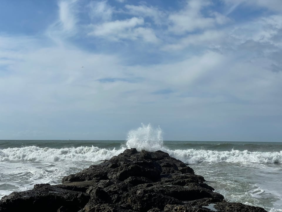 A breaker made of rocks extends into the ocean. Waves churn and a big wave crashes straight up at the end of the breaker
