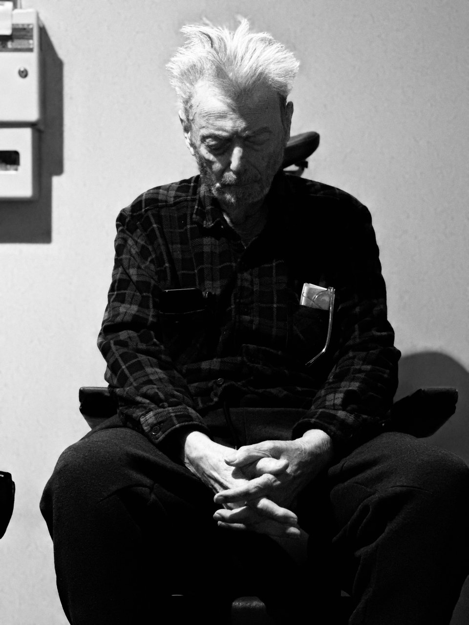 b/w photo of elderly white man sitting in an exam chair at an ophthalmologist's office. His fingers are interlaced, his head is down, and he has a shock of white hair.