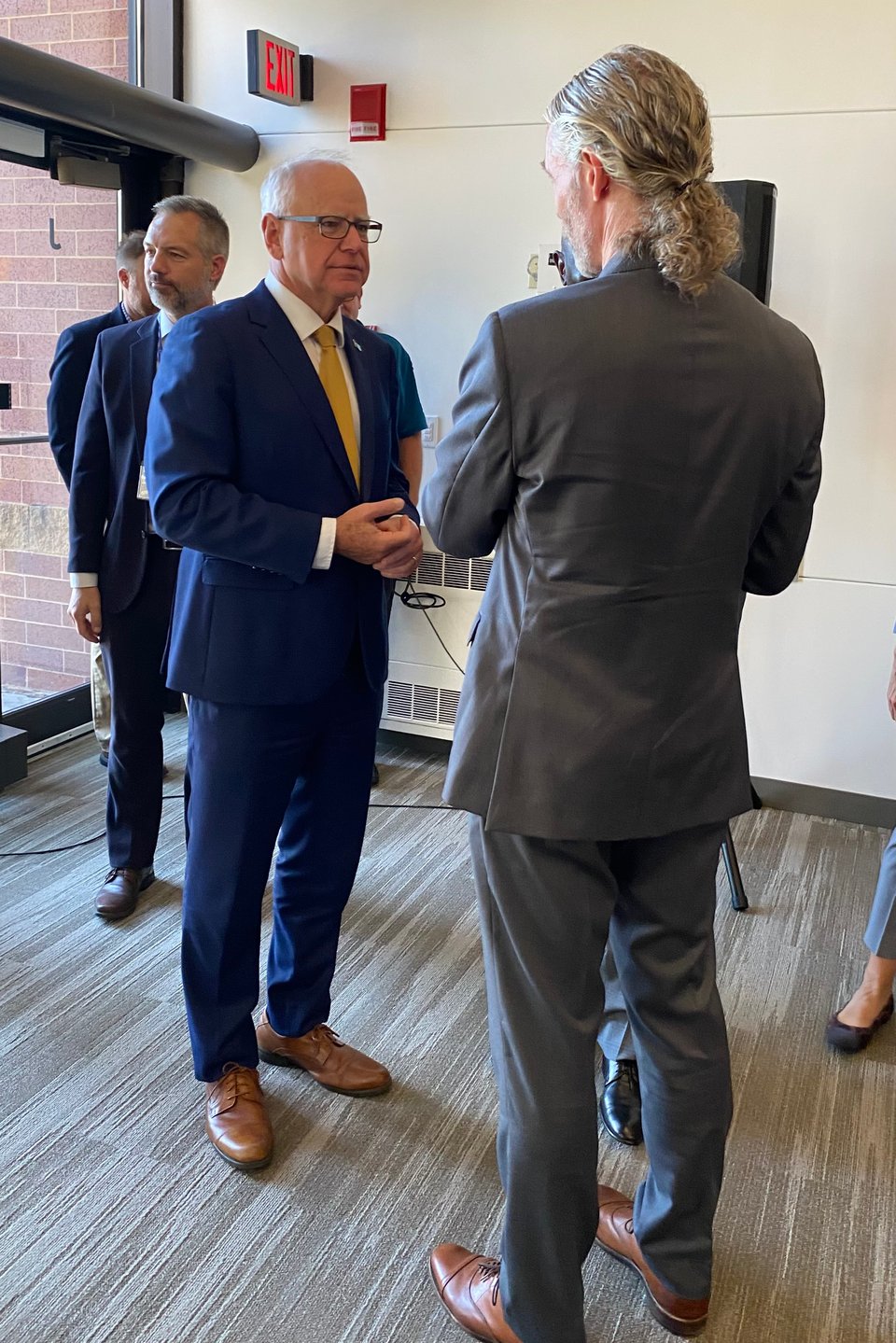 photo of MN Gov. Walz in a blue suit talking to a white man in a gray suit; the other guy has his back to the camera mostly, and has a long curly ponytail