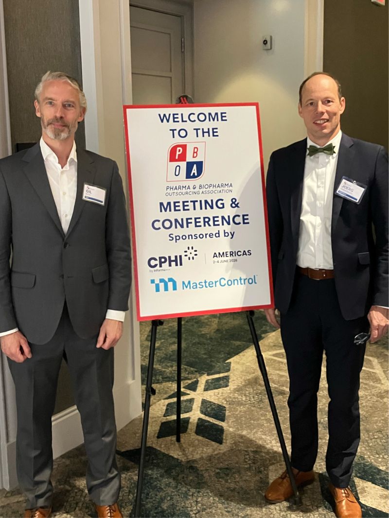 photo of two white men standing beside an easel with a poster welcoming people to a conference. The guy on the left is in a gray suit and white dress shirt with no tie, and the guy on the right is in a navy suit, white dress shirt, and green & blue bow-tie. They're both smiling-ish.