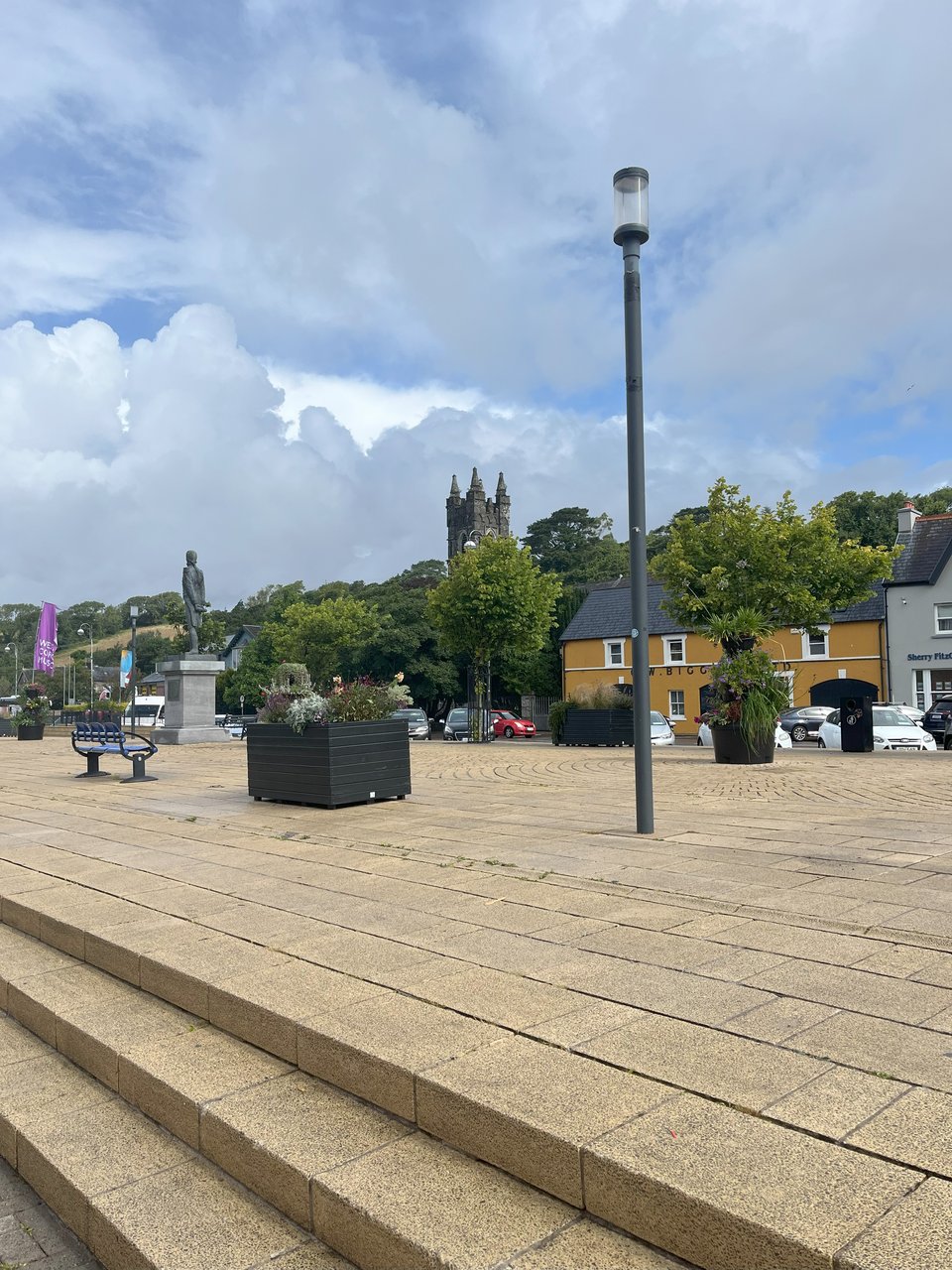 A photograph of Wolfe Tone Square in Bantry. The white clouds are fluffy and in high contrast to the peaks of blue sky. Most of the foreground is taken up by the sand coloured bricks of the square. In the distance is a church steeple, shrouded by vivid green trees. Dotted across the square are boxes of flowers, public benches, rubbish bins, and street lamps. To the left is a statue on a plinth. To the right, cars are parked in front of a mustard yellow two storey building.