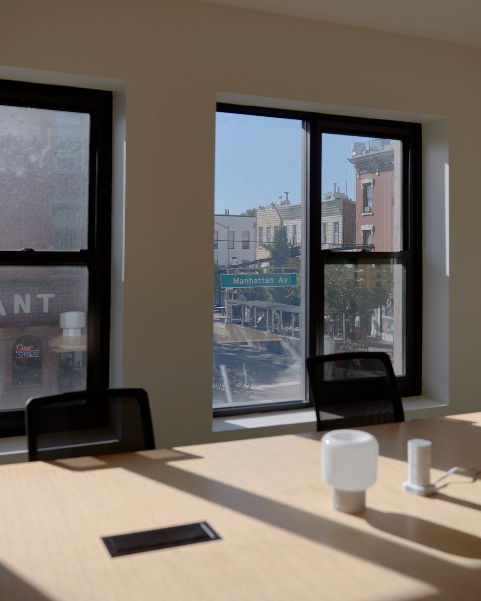 A long work table and desk chairs before a window looking out on the street. The street sign for Manhattan Av can be seen through the window.