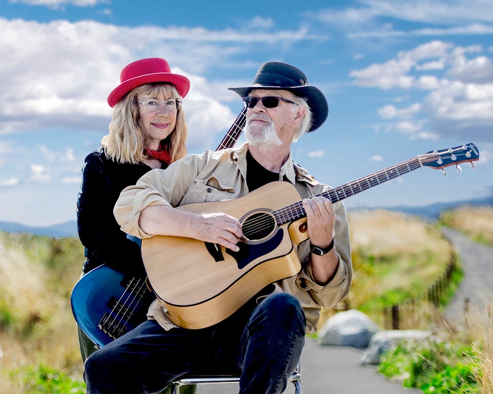 a woman n a hat standing behind a man with a guitar seated on a chair outdoors