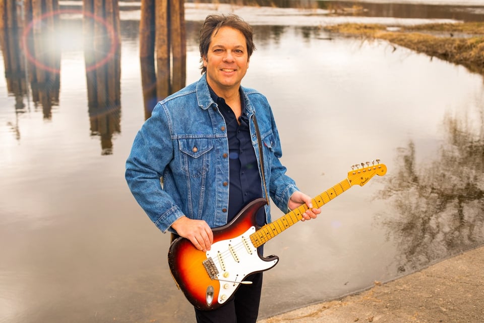 A man in a jean jacket holding a tobacco-burst Fender Stratocaster guitar stands in front of a pond.