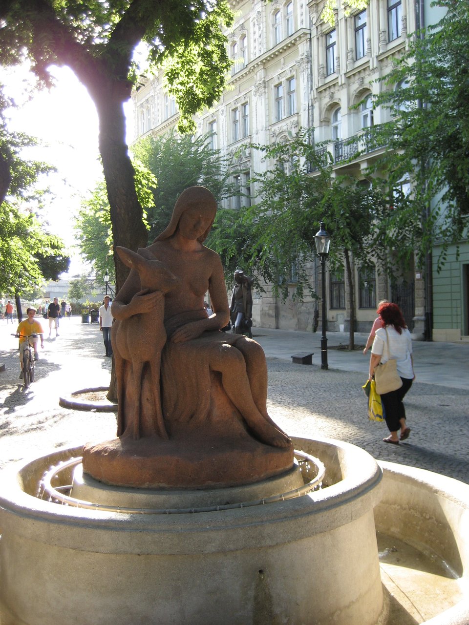 A sculpture of a seated woman wearing a dress, with long hair. Her right arm is gently embracing and holding a small deer.