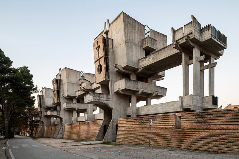 A brutalist building that consists of three square-shaped structures boosted about 20 feet off the ground on concrete pillars, with a concrete walkway connecting them, and square concrete verandas sticking out from their sides. The concrete is grey and weathered, with black streaks