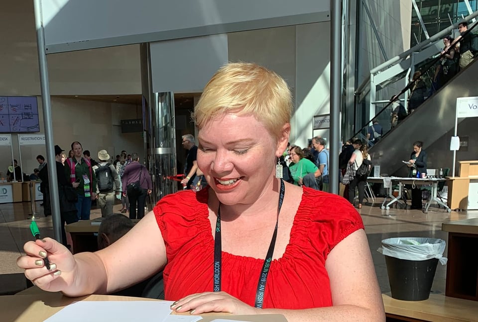 Kate wearing a red peasant blouse, a convention lanyard, a blonde pixie cut, and a big grin. She is holding a pen as she prepares to sign a document. In the background, you can see a busy convention centre foyer.