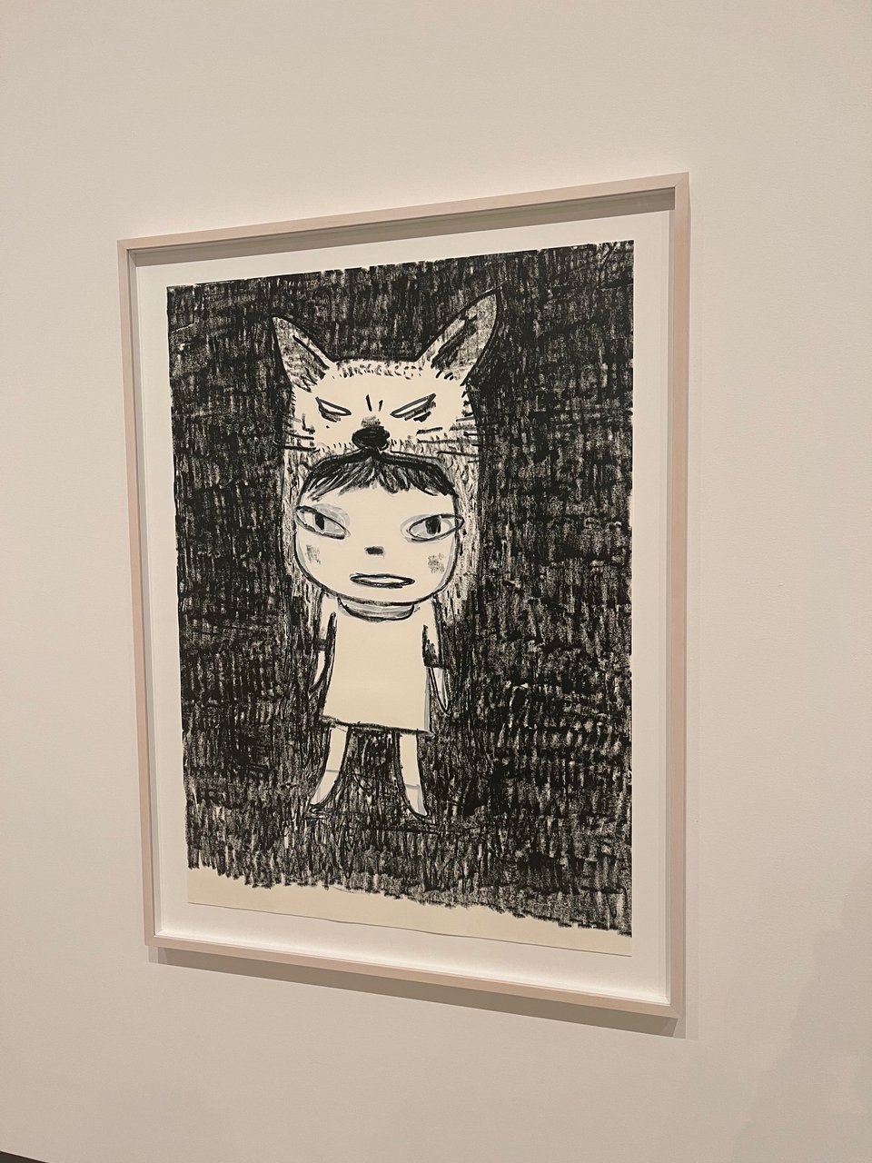 A photograph of a Yoshitomo Nara illustration. It is a monochrome piece, black and white, using a paint marker. The background has been coloured black. In the middle of the canvas is a drawing of a child in a long t-shirt or dress. They are wearing a cat or fox's head as a hat, and some of their fringe pokes out. They look head on at the viewer.