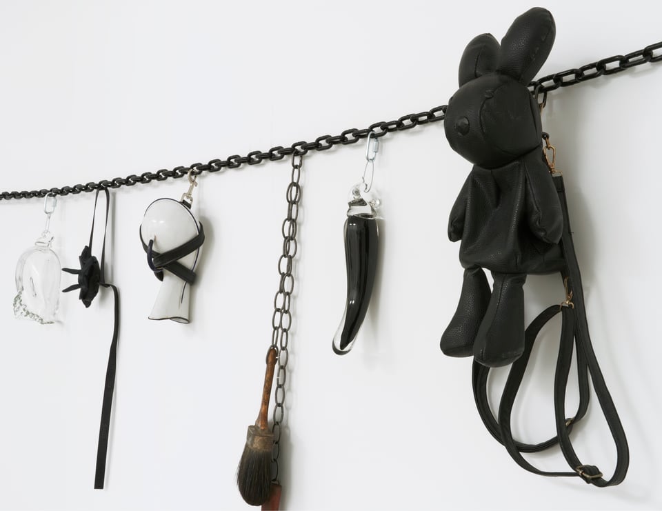 Detail of an artwork by Nayland Blake showing a black chain strung horizontally on a white wall. Various "charms" are clipped to it, seriously and comically referencing leather-fetish and BDSM practices. Including restraints and a black leather backpack shaped like a bunny.