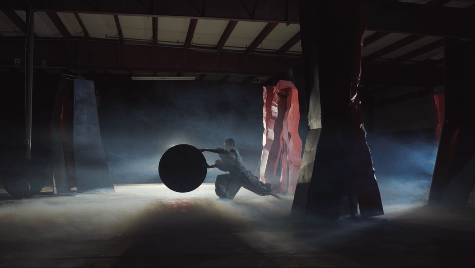 A dancer in loose, white clothing holds a circle with arms extended, inside a warehouse populated by various sculptures and shapes. The space is dark save for a diffuse light and fog, giving it a mysterious, theatrical atmosphere.
