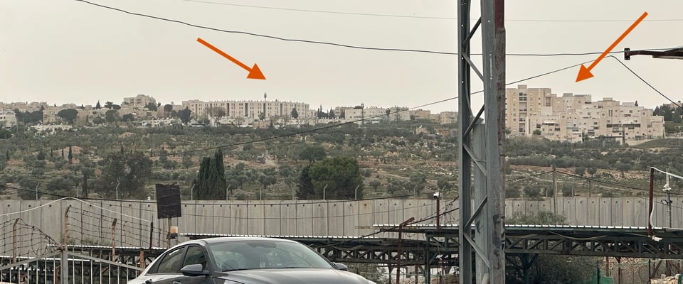 Behind a car, a long wall of tall concrete slabs. In the distance behind that, two collections of buildings forming a small city