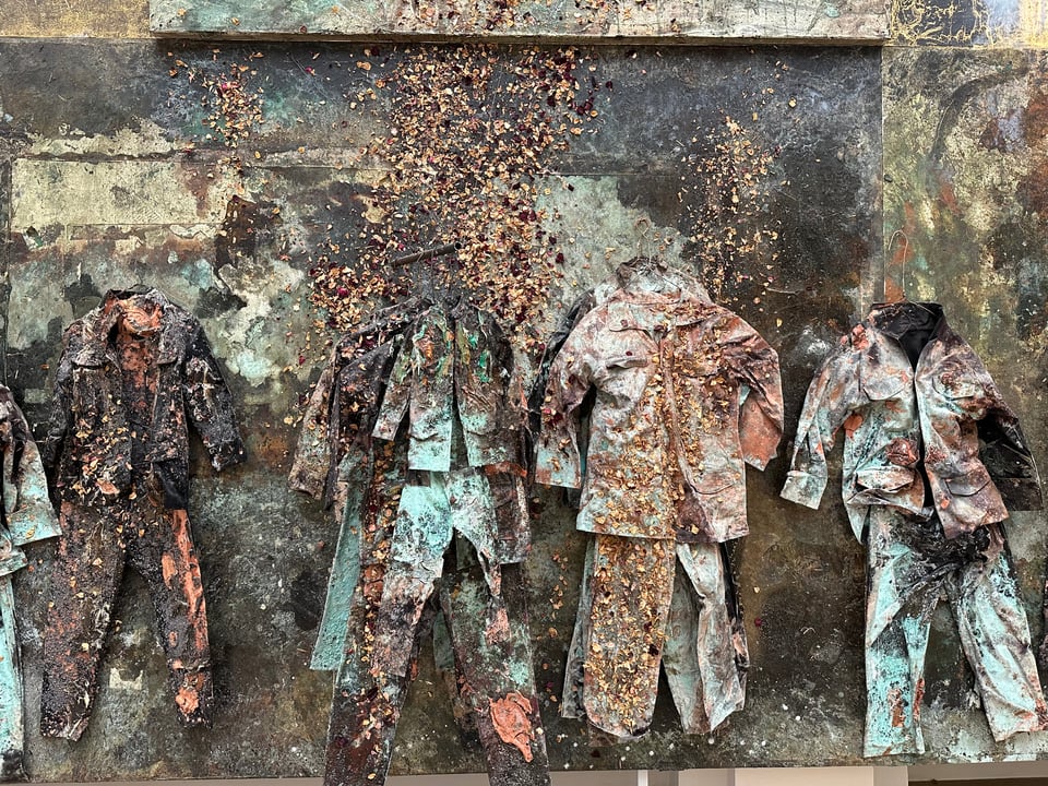 detail of Anselm Kiefer's "Sag mir wo die Blumen sind", showing a series of work clothes painted stiff, hanging against a painted canvas backdrop, with flower petals stuck to them
