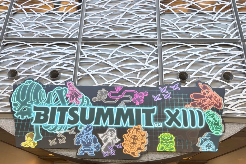 A photo of a large sign above a crowded convention. The sign has various Japanese demons drawn in neon lights. Text reads, "BitSummit XIII”