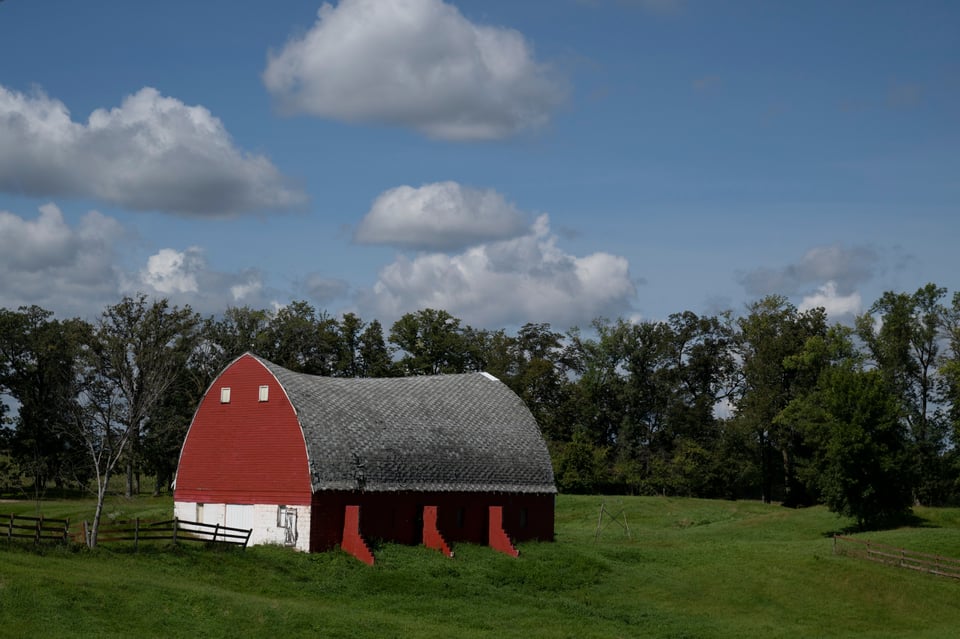 A red barn with an arched pointed roof and buttresses, in a green field backed by trees.