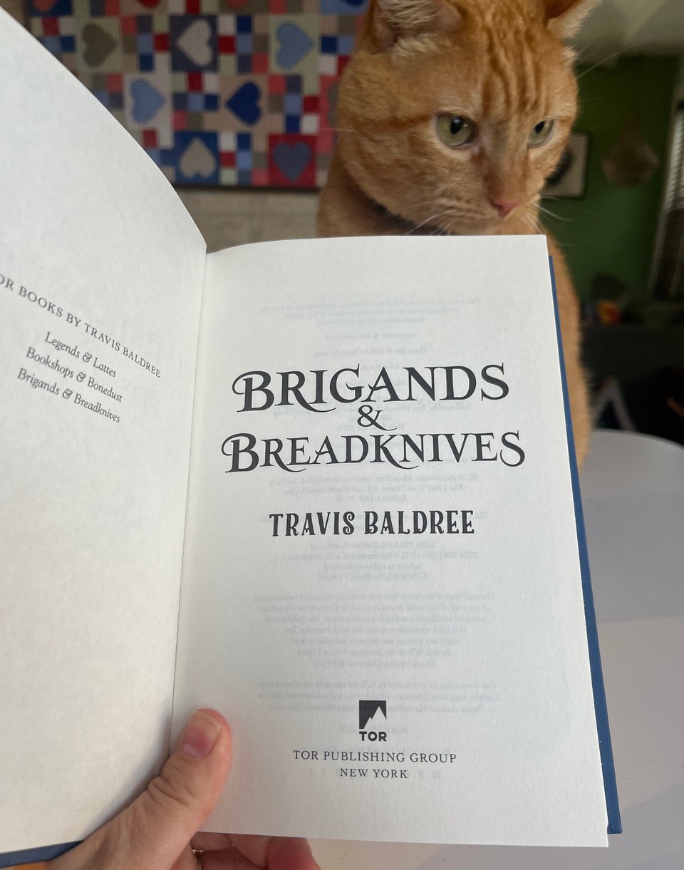 A white person's hand holds a book open to the title page, with an orange tabby cat in the background.