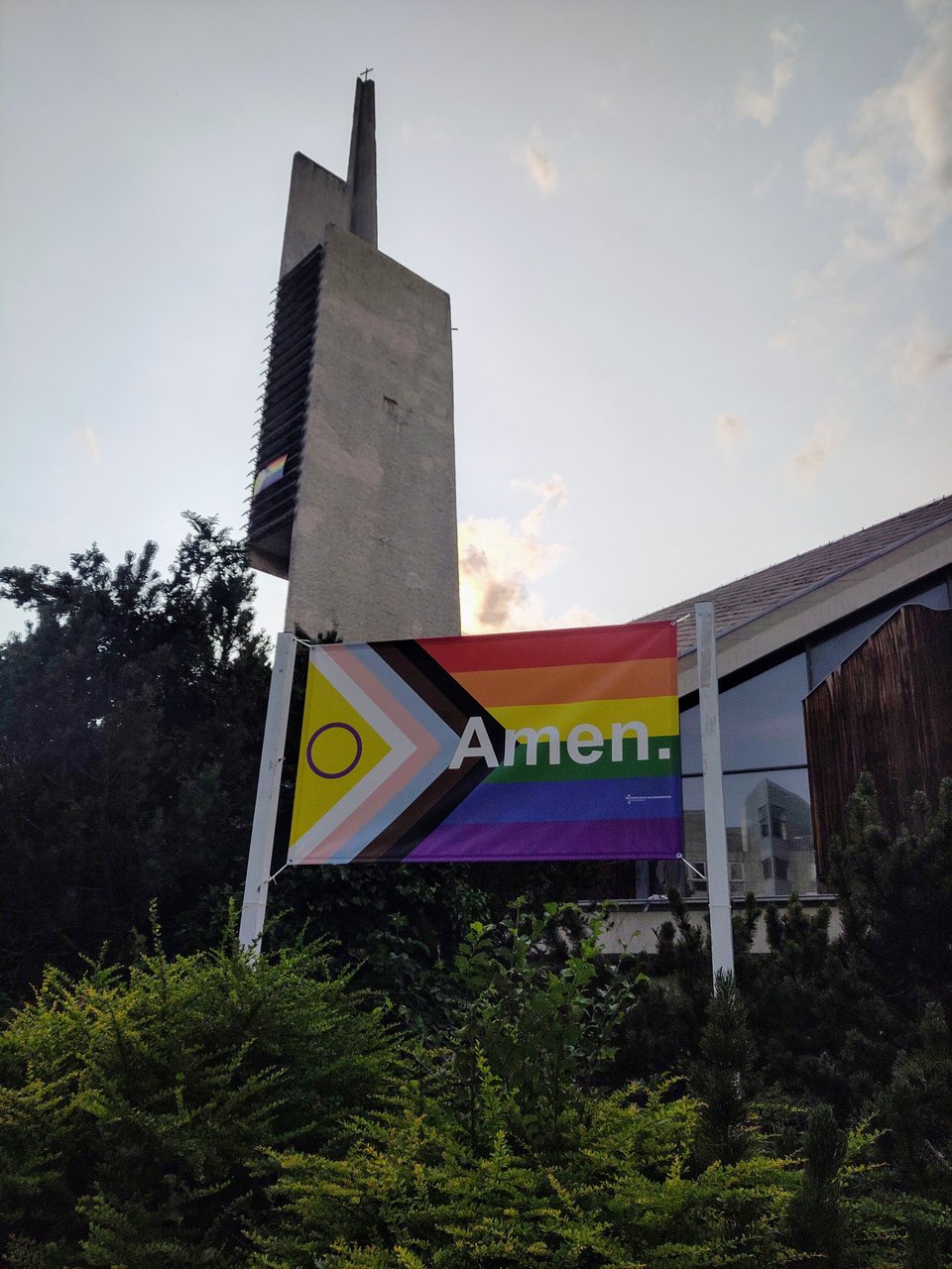 Brutalistischer Kirchturm, davor eine große Progress-Pride-Flagge mit dem Aufdruck "Amen."