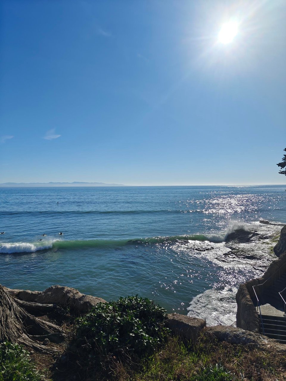 view from a cliff overlooking the Pacific ocean on a clear, sunny day. a wave is just breaking rather dramatically, right behind a staircase that leads to the beach at the foot of the cliff. there are some tree roots visible to the left in the foreground.
