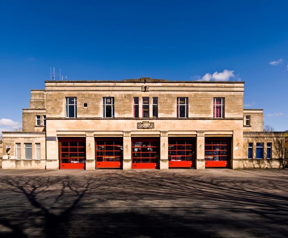 A 1930s fire station. It has five bay doors set into a wide front. Above that is a set of narrower windows that are set back. To either side of the main frontage are smaller rooms set back a little. The whole is clad in creamy Bath stone.