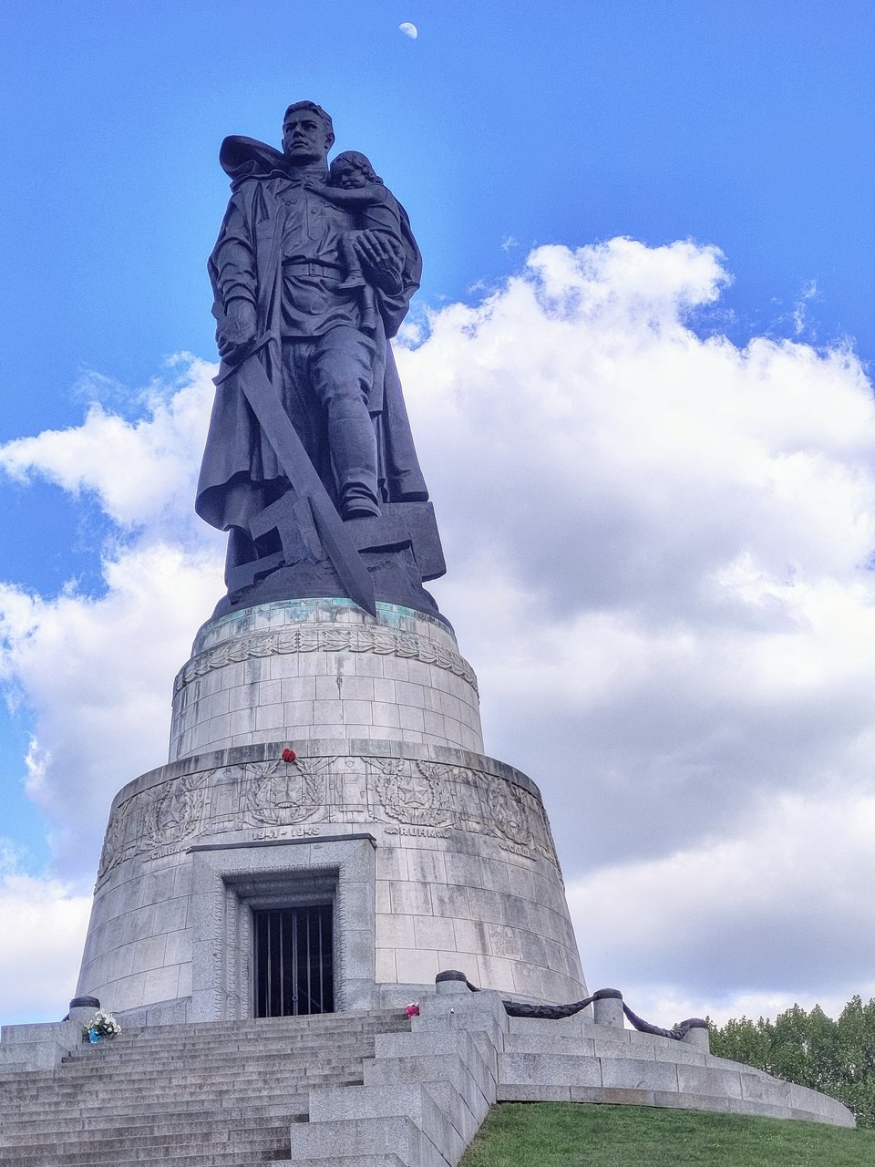 Sehr hohe Statue eines Soldaten, der im linken Arm ein kleines Kind trägt, in der rechten Hand ein Schwert hält, dessen Spitze auf ein am Boden zertrümmert liegendes Hakenkreuz zeigt. Am oberen Bildrand ist im blauen Himmel der Mond zu erkennen.