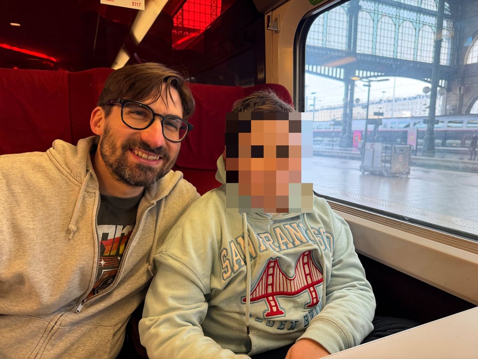 A man and a young boy sit in chairs on a train. The young boy's face is blurred.