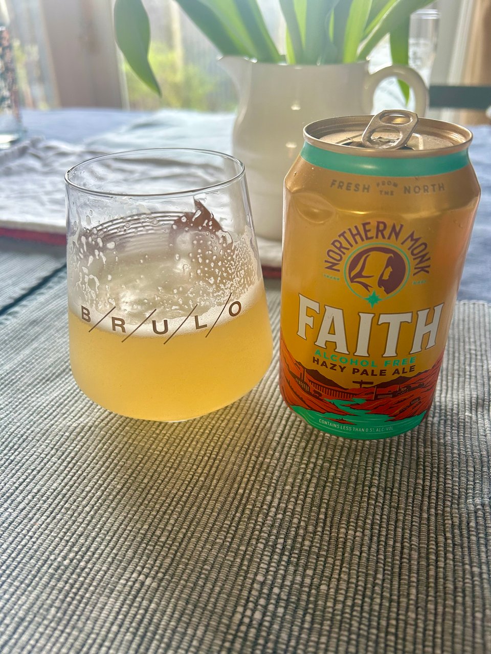 A glass with hazy beer in it sits next to its can with Northern Monk branding on a textured dark blue and white tablecloth. There's a cream jug with apple green tulip stems behind. Image by Rowan Ambrose.