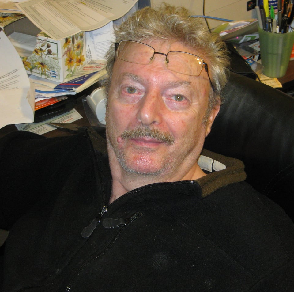 photo of white man in black zip-up track shirt, with a mustache, and reading glasses on his forehead