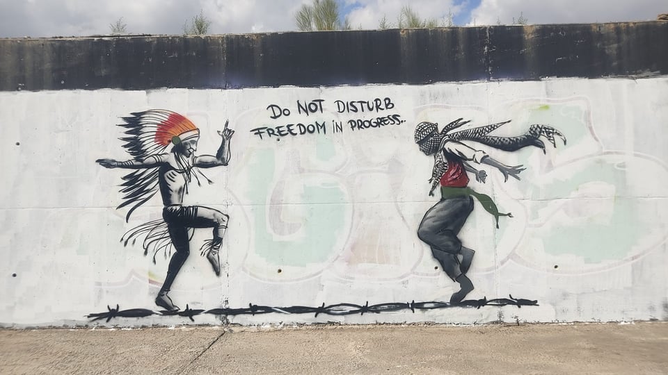 Graffiti on a wall with a white background, showing an indigenous person and a Palestinian dancing facing each other on a strand of barbed wire with the words "Do Not Disturb Freedom in Progress" between them