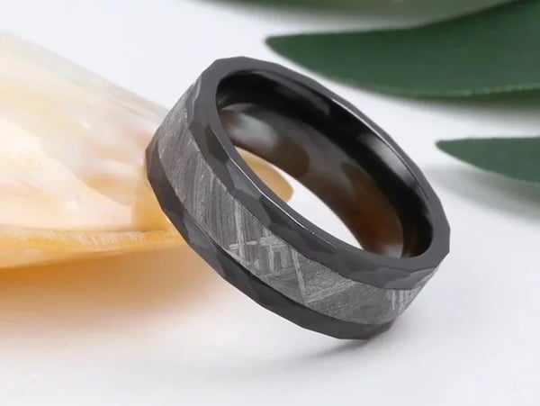 Un anillo con tres franjas. Las dos exteriores están trabajadas en patrones geométricso regulares con algún tipo de material negro y la franja central, más ámplia es metálica y tiene un patrón de líneas diagonales característico de los meteoritos metálicos sometidos a presión y a altas temperaturas.