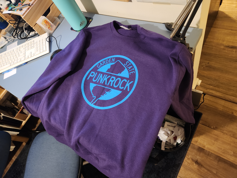 A photo of a purple sweatshirt with the Garden State Punk Rock graphic printed on it in blue.