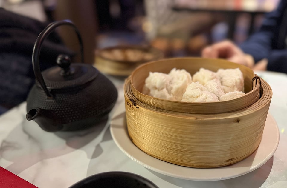 A bamboo steamer filled with three dim-sum dumplings on a table. Next to it is a metal teapot.