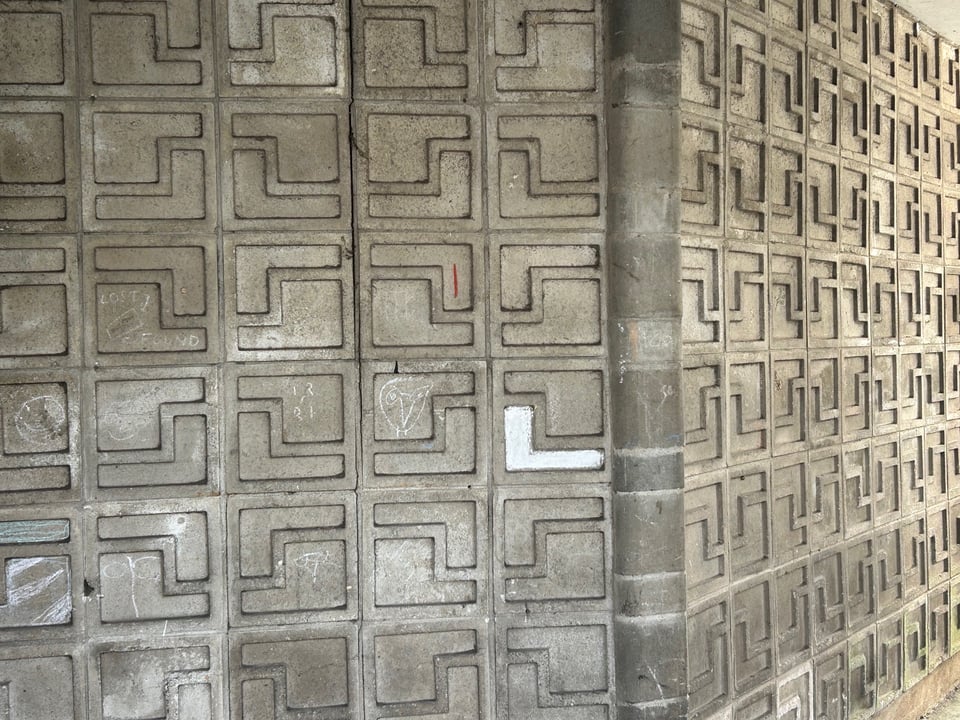 A bas relief wall made up of squares of cast concrete that have been put together to resemble interlocked plus signs (or crosses). Someone has coloured a bit of one in white.