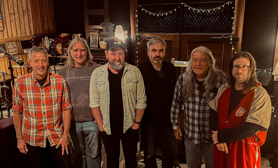 Six men stand in a rough line in a recording studio where they made a record.