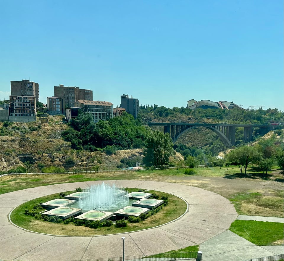 a fountain in the foreground, in front of a gorge, with a bridge spanning it, and buildings in the background