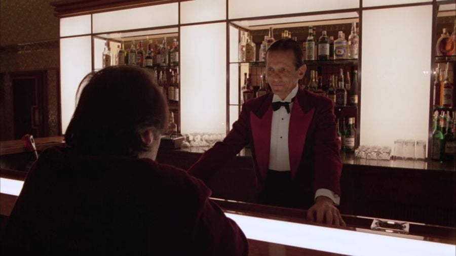 Jack Torrance sits at a bar. The barkeeper wears a red jacket, white shirt, and a black bow tie. The shelves behind him are filled with liquor bottles.