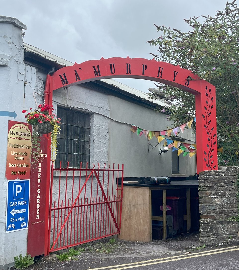 A photograph of the back entry to Ma Murphy's, a pub in bantry. The sky overhead is grey and overcast. To the right is a glimpse of an old stone wall. To the left are signs on a white brick wall. One sign is blue and directs people to the car park. The sign above it is orange, yellow, and red, and it lists out the amenities in Ma Murphy's. The red iron gate is wide and open, allowing people into the beer garden. An archway, painted the same red, reads "Ma Murphy's". A hanging basket full of red flowers hangs from the left. Colourful bunting can be seen leading down the alley.