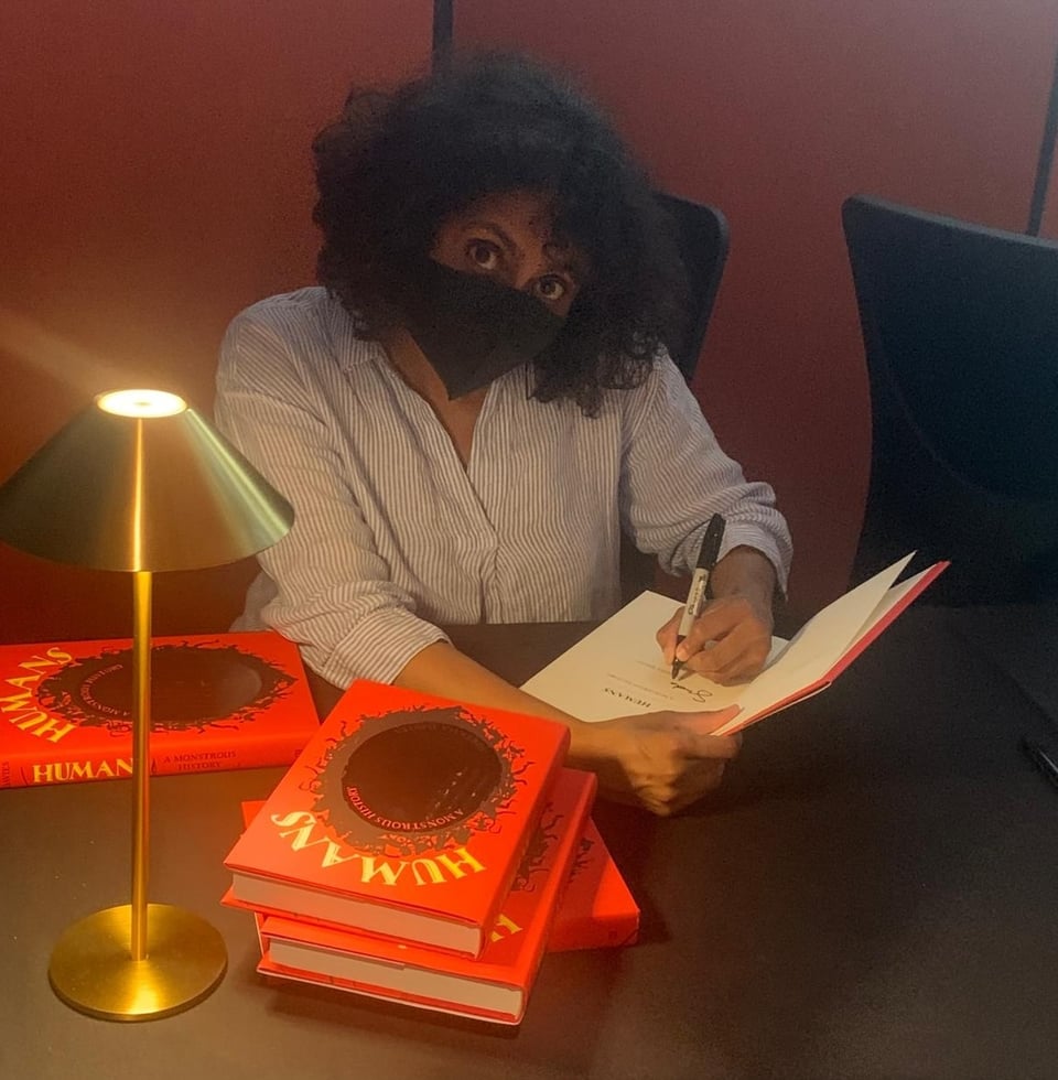 Dark-haired woman in a black face mask looks up quizzically while signing a pile of copies of “Humans: A Monstrous History.” She sits at a desk covered in a black cloth. On the left is a brass-coloured desk lamp with a triangular shade.