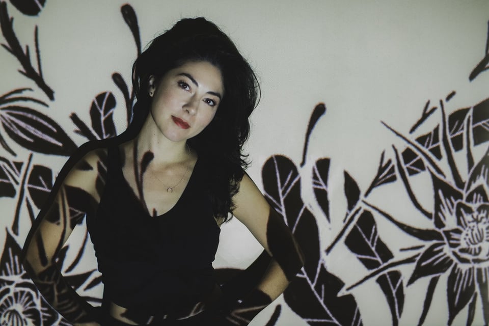 A woman in her 30s with long dark hair stands with her hands on her hips, head tilted to the left, looking at the camera. Drawings of leaves and flowers are projected onto her.