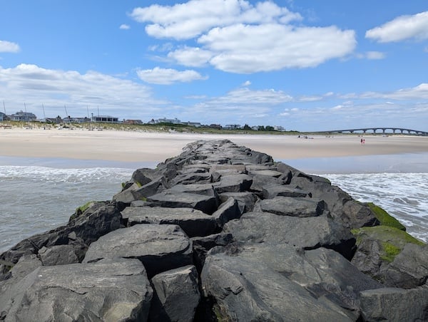 A rocky promontory extending back from the ocean onto a beach, with dunes, houses and a causeway in the distance