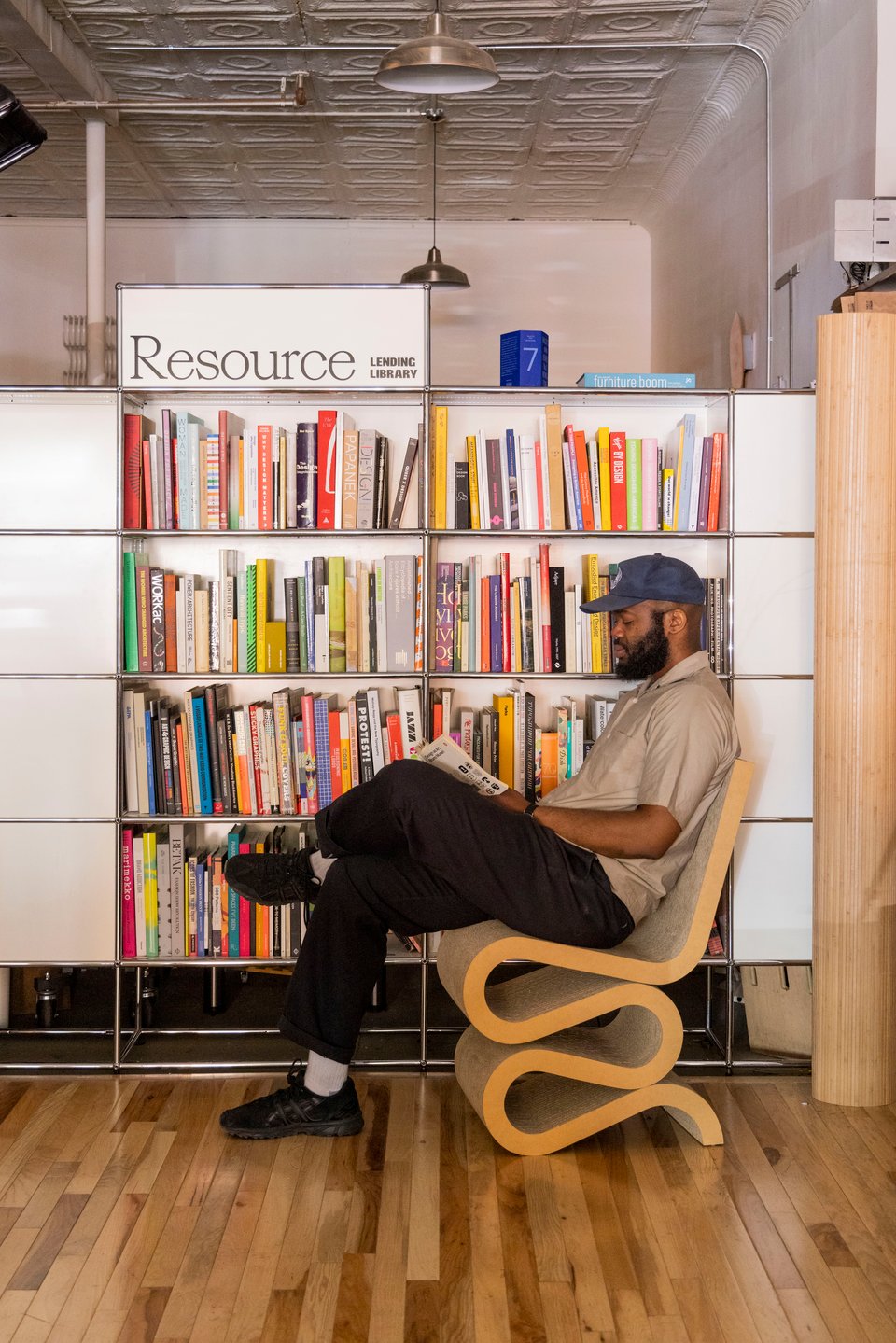 A person sits on a nicely designed chair with a squiggly base reading a book from the Resource Library shelves at Lichen.