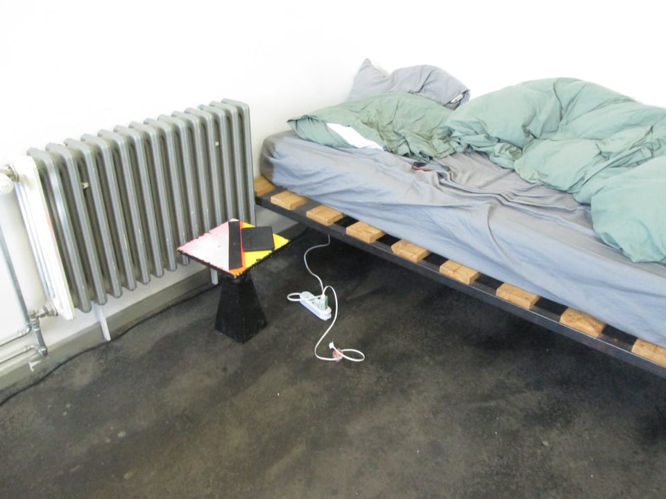 a stark image of a disheveled bed, nightstand, radiator and cables