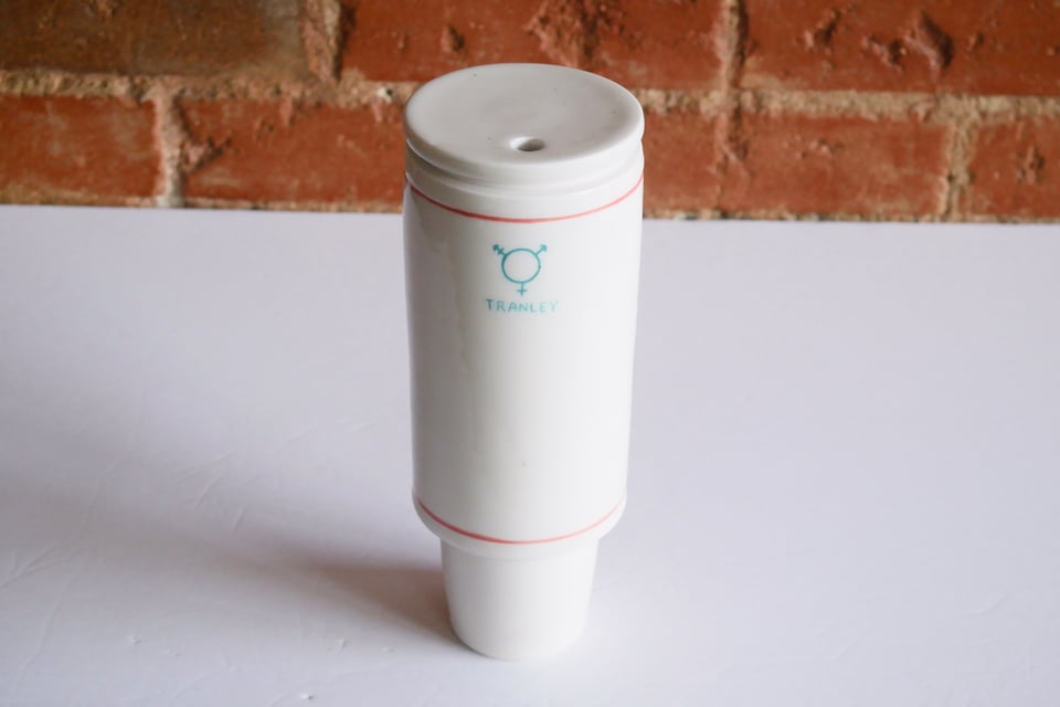 The classic stanley cup form, with a car cup-holder friendly base and wider top - but this one is a white porcelain, with subtle pink and blue decorations and a three-pronged transgender symbol with the word TRANLEY written underneath