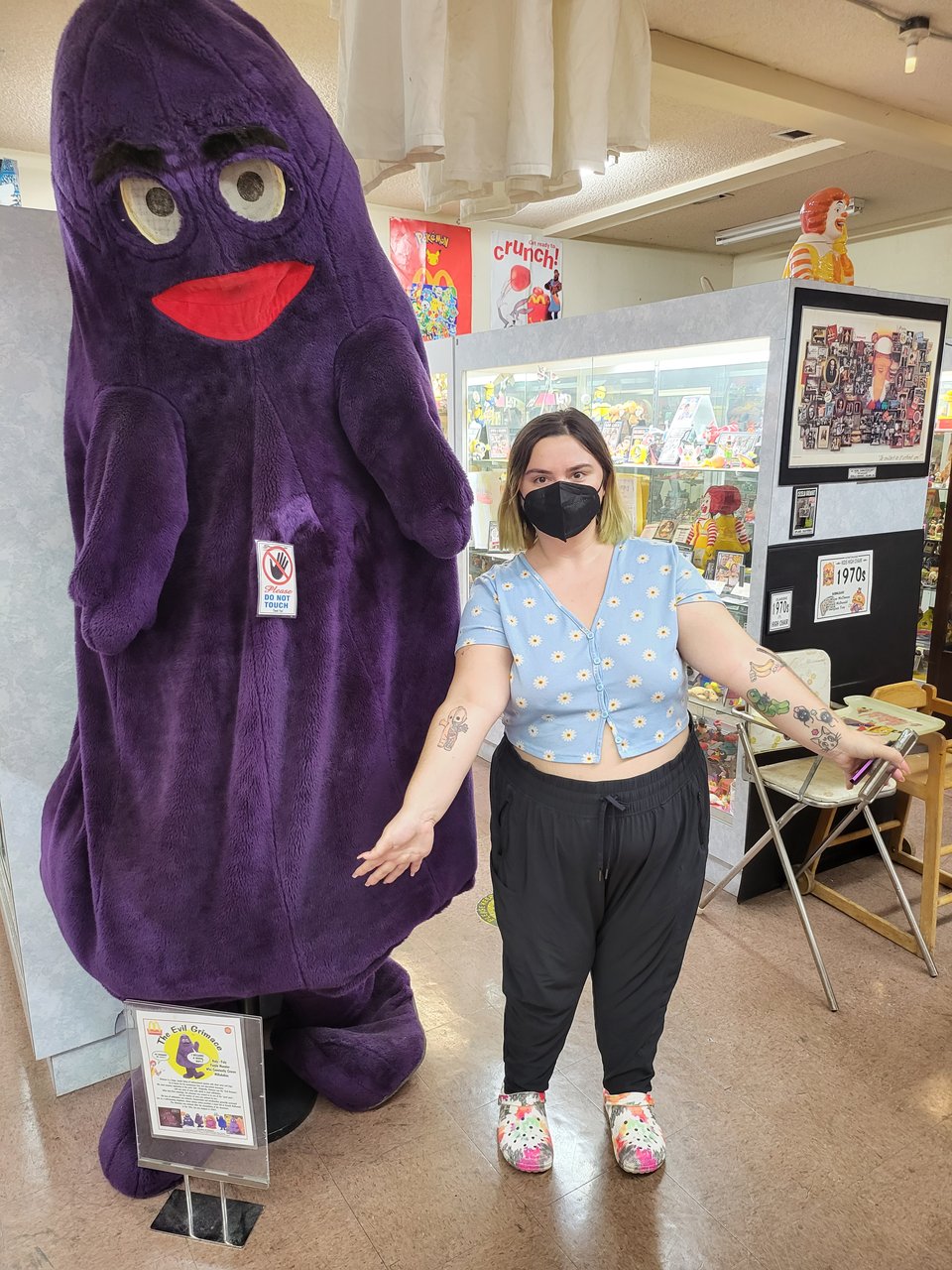 Becca stands with their arms spread in the Original McDonald's Museum to mimic an empty Grimace, the big purple whatever he is monster, costume on display.
