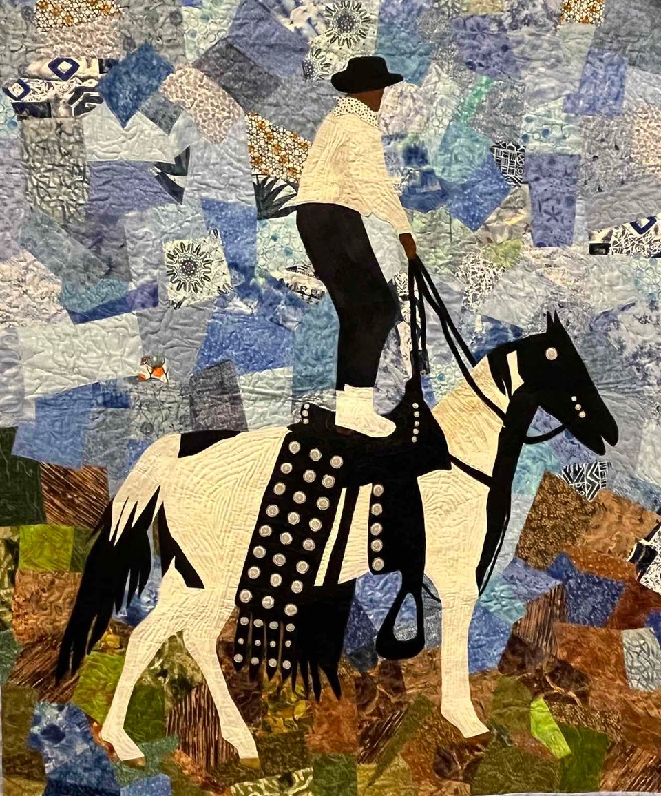 quilt depicting a person with dark brown skin, a white long sleeved jacket, black pants, and white boots standing on the back of a black and white horse and holding black reins. the sky is a patchwork of pattered blue fabrics and the ground is a similar patchwork of patterned fabrics in earth tones