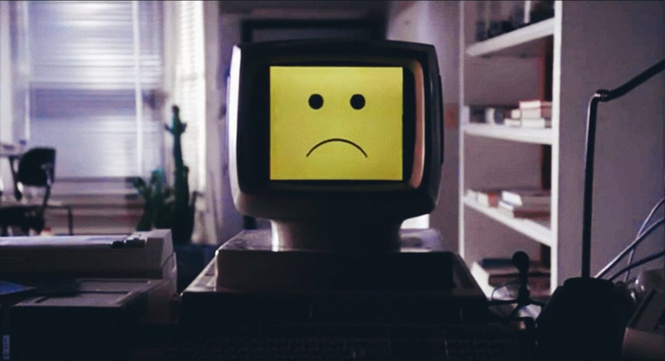 A frame from Electric Dreams (1984). A computer with a yellow screen and a frowney face.
