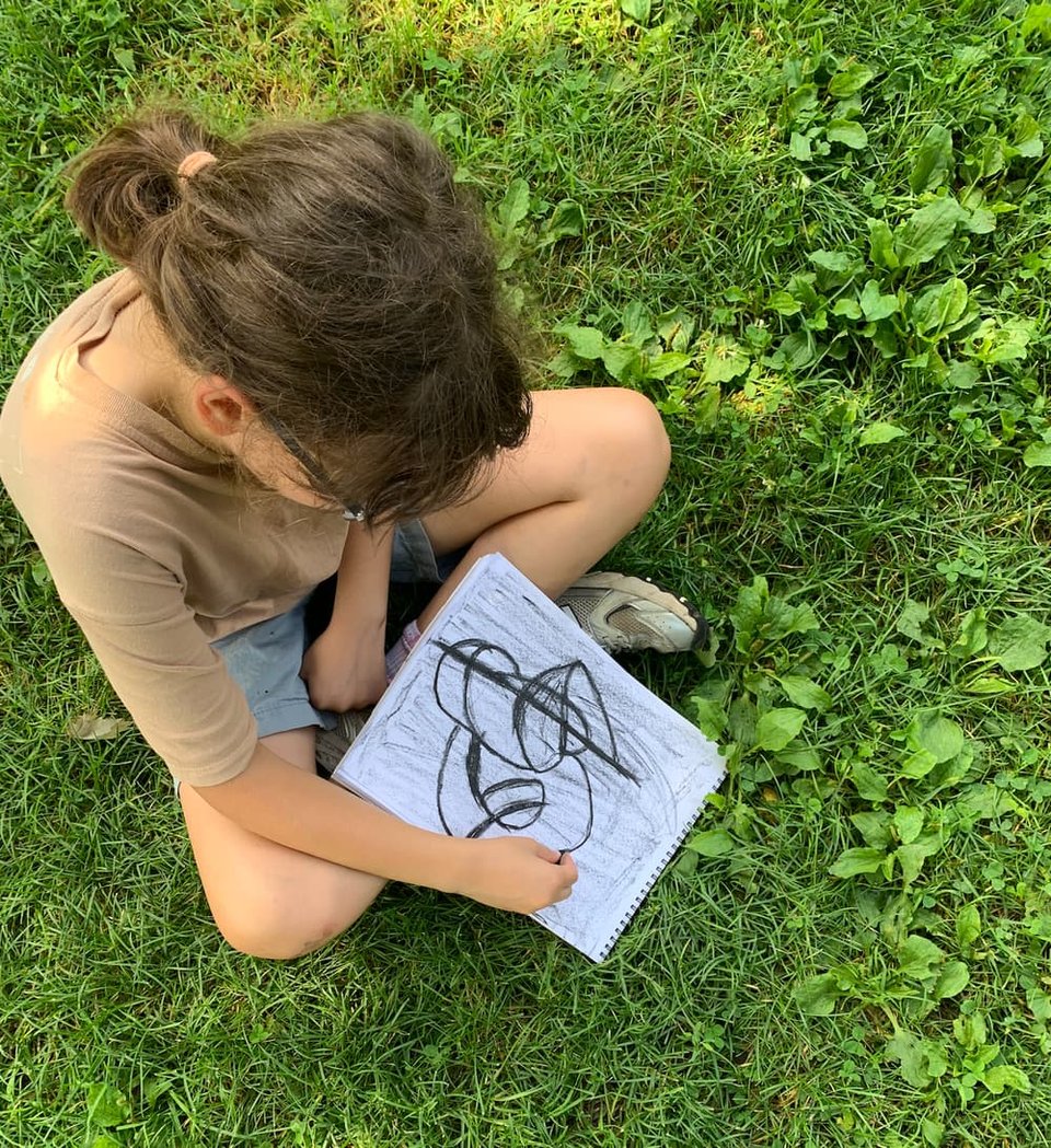 Photograph of a child, seen from above, drawing in a sketchbook on the grass