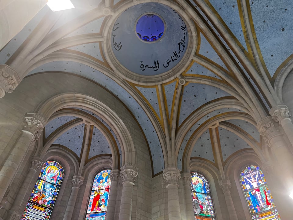 A vaulted church ceiling with a dome containing writing in Arabic, and below that, stained glass windows
