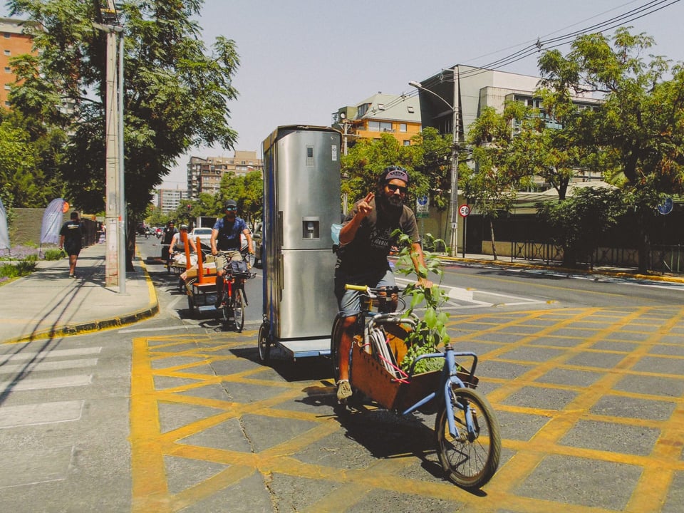 Several people carrying objects on cargo bikes. The person in front has a fridge on their cargo bike trailer, and a plant on the front.