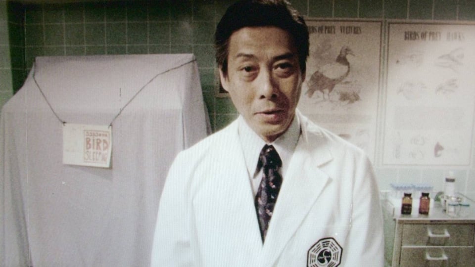 An image of the Dharma initiative. An Asian man with a tie wearing a white lab coat with a stylized hexagon symbol on the breast stands in front of some mysteroius bird-related charts, plus a box with a white sheet over it and a label that says BIRD.