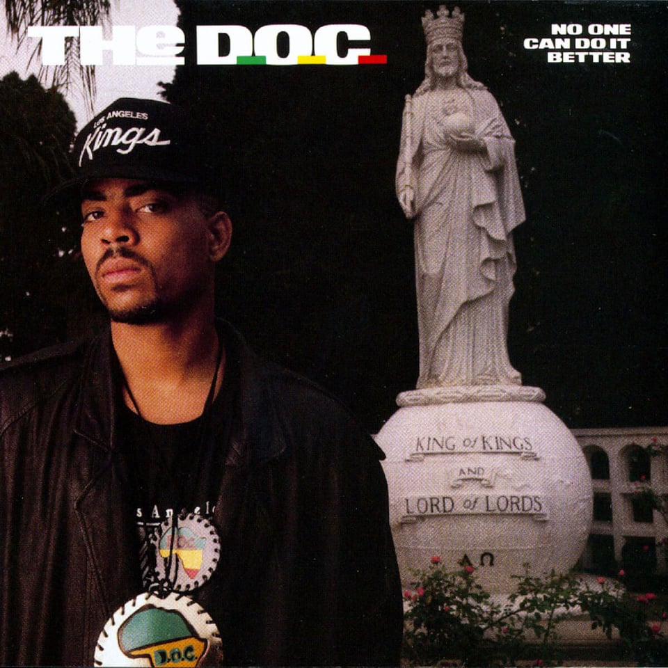 A color photograph of The D.O.C., a young Black man wearing a leather jacket, a black Sacramento Kings cap, and two medallions, possibly made of leather, showing Africa. He has a wary, suspicious look on his face. He is outside, possibly in a park somewhere. Behind him is a statue of I'm guessing Jesus, but wearing a crown, holding an orb, and a staff in the other hand. He's is on an orb-shaped pedastal that reads "KING OF KINGS AND LORD OF LORDS."